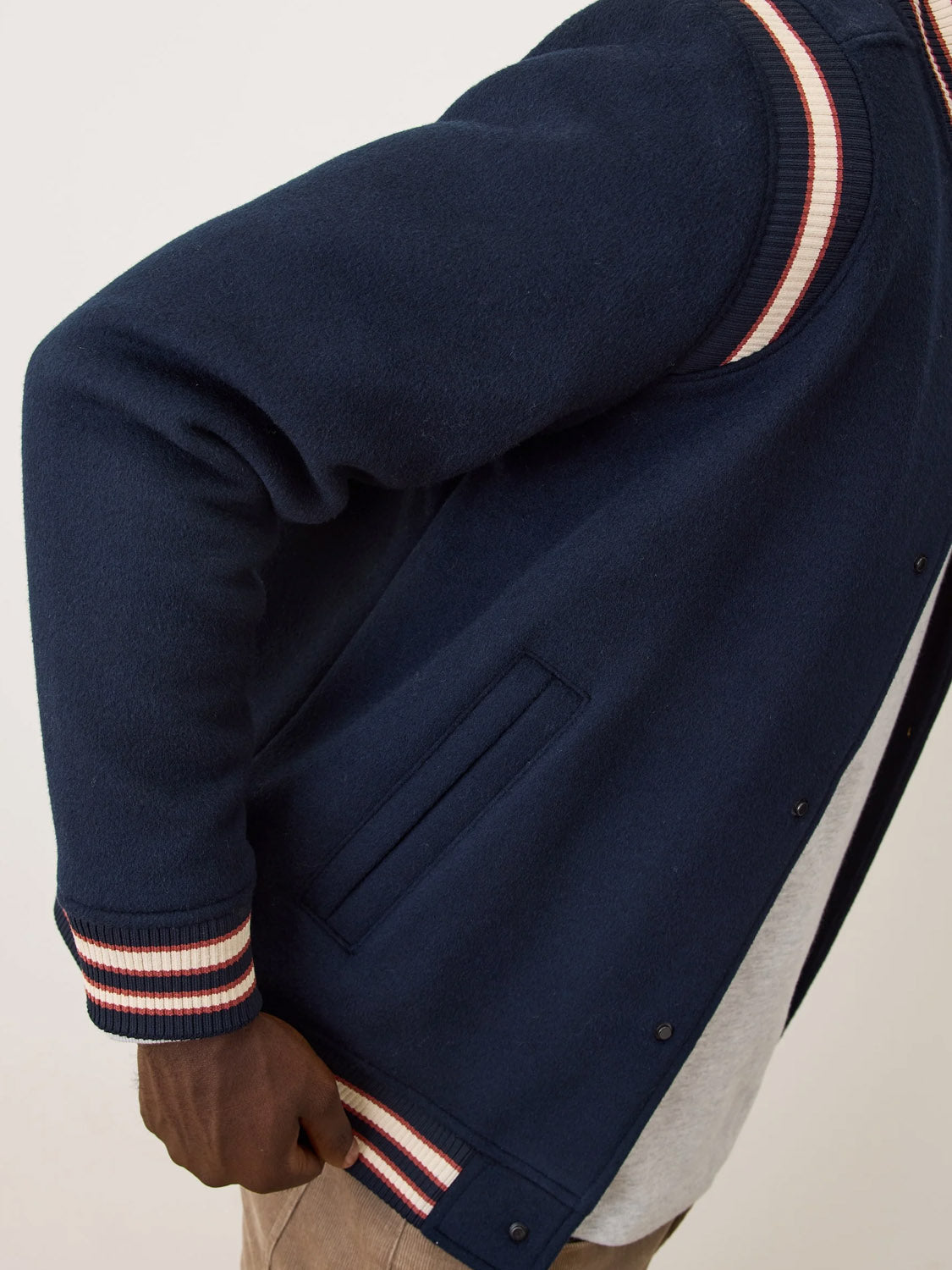 A person stands in profile with one hand in their pocket, wearing the Marine Layer Fields Wool Varsity Jacket in navy blue, featuring red, white, and blue striped cuffs and collar.