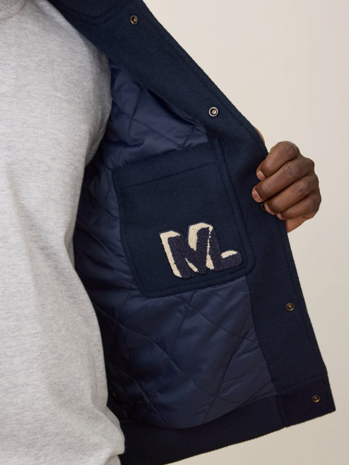 A person opens the Marine Layer Fields Wool Varsity Jacket in navy, revealing an inside pocket with an abstract white and navy patch. They are wearing a light grey shirt underneath.