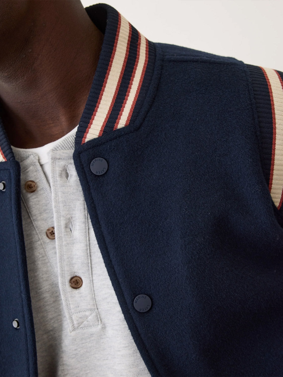Close-up of a person wearing the Marine Layer Fields Wool Varsity Jacket, featuring a red and white striped collar, layered over a light gray henley. Only part of the face and upper body is visible.