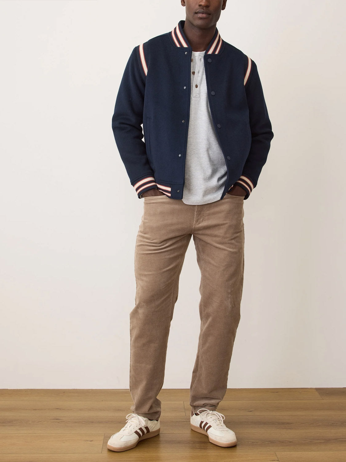 A man stands indoors wearing the Marine Layer Fields Wool Varsity Jacket, featuring striped cuffs and collar, paired with a light gray henley shirt, tan corduroy pants, and white sneakers with brown stripes.