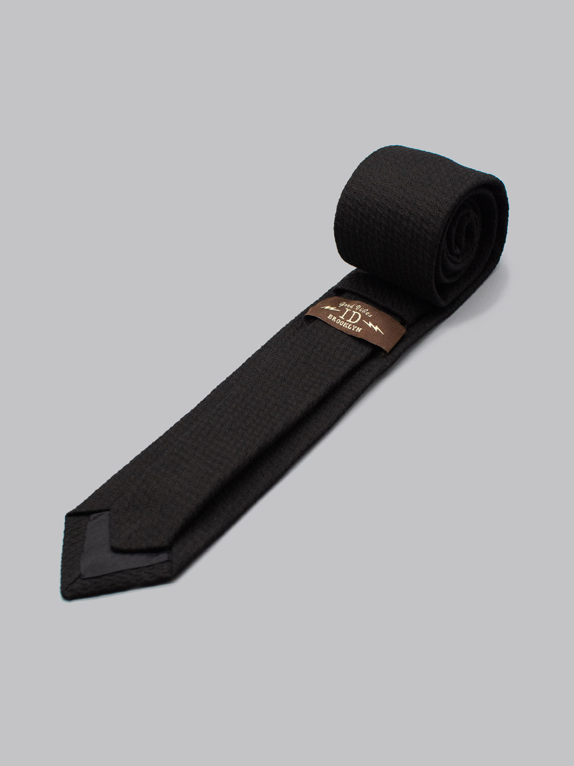 The ID Cotton Tie Venice by ID Menswear is a neatly rolled black cotton tie with a subtle textured pattern, displayed on a gray surface. A brown Handmade in Brooklyn label is sewn underneath—ideal for business formal occasions.