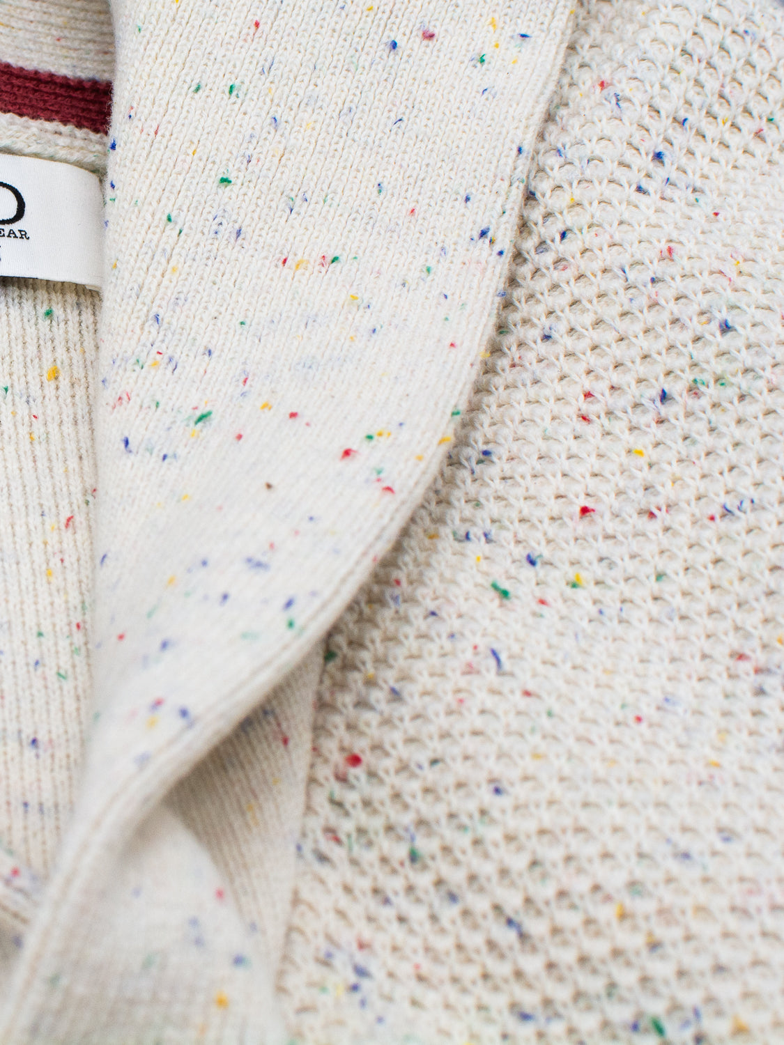 Close-up of cream-colored textured fabric with multicolored specks, showcasing a knit pattern with red, blue, and yellow. The visible label reads ID Menswear, echoing the speckled yarn used in crafting the Vermont Shawl Collar Cardigan.