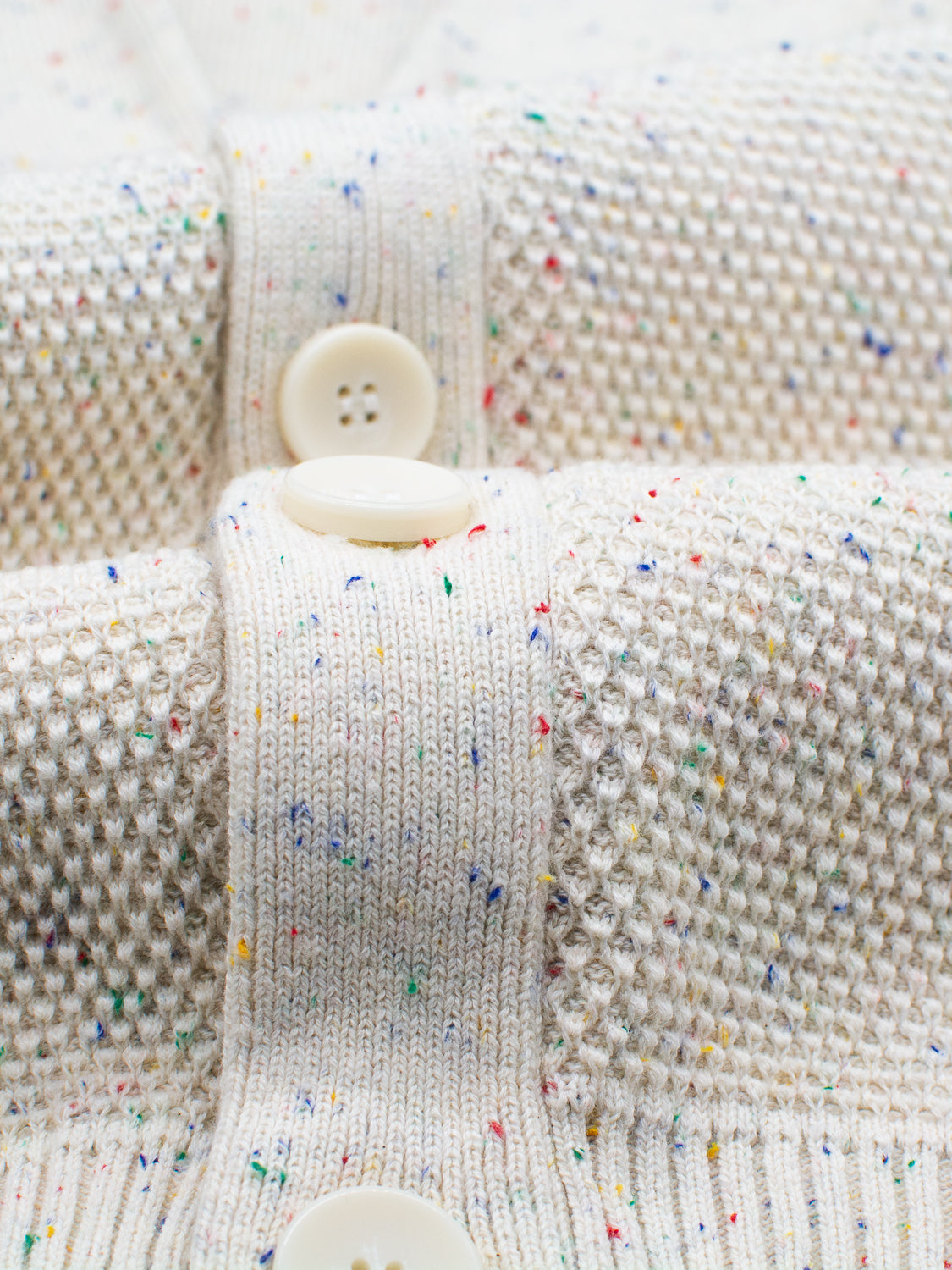 A close-up of the ID Menswear Vermont Shawl Collar Cardigan reveals a cream-colored design made from speckled yarn with multicolored flecks. It features a stylish shawl collar, a mix of ribbed and textured knit patterns, and three white buttons adding intricate detail to the cozy garment.