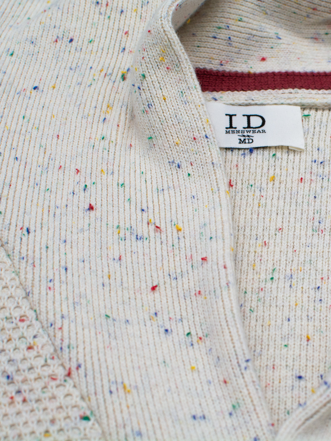 Close-up of a cream-colored Vermont Shawl Collar Cardigan by ID Menswear, made with multicolored speckled yarn. It features an elegant shawl collar, ribbed and mesh textures, and a label reading I.D. MENSWEAR MD sewn into the neckline.