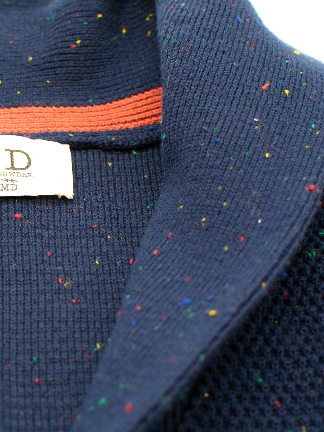 Close-up of the Vermont Shawl Collar Cardigan by ID Menswear, featuring navy blue textured knit with colorful specks and an orange stripe near the shawl collar. A clothing label can be seen inside.
