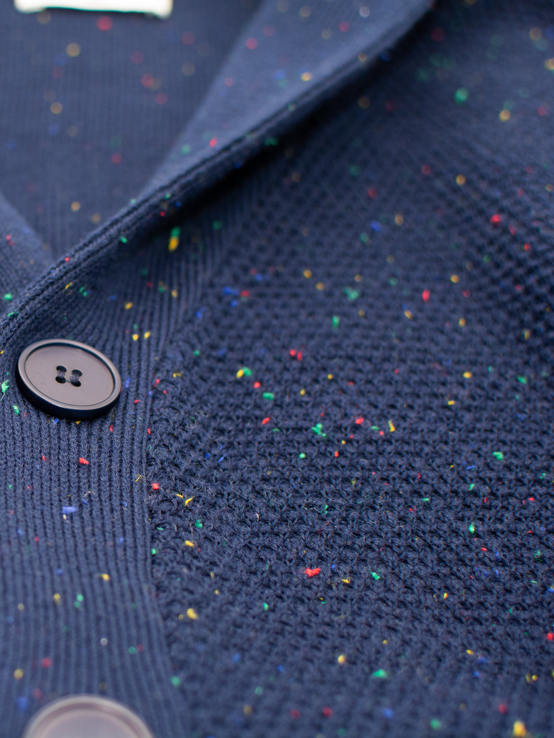 The Vermont Shawl Collar Cardigan by ID Menswear is highlighted in a close-up, showcasing speckled navy blue yarn and black buttons. With smooth and ribbed textures, it exudes a cozy vibe that is accentuated by the stylish shawl collar.