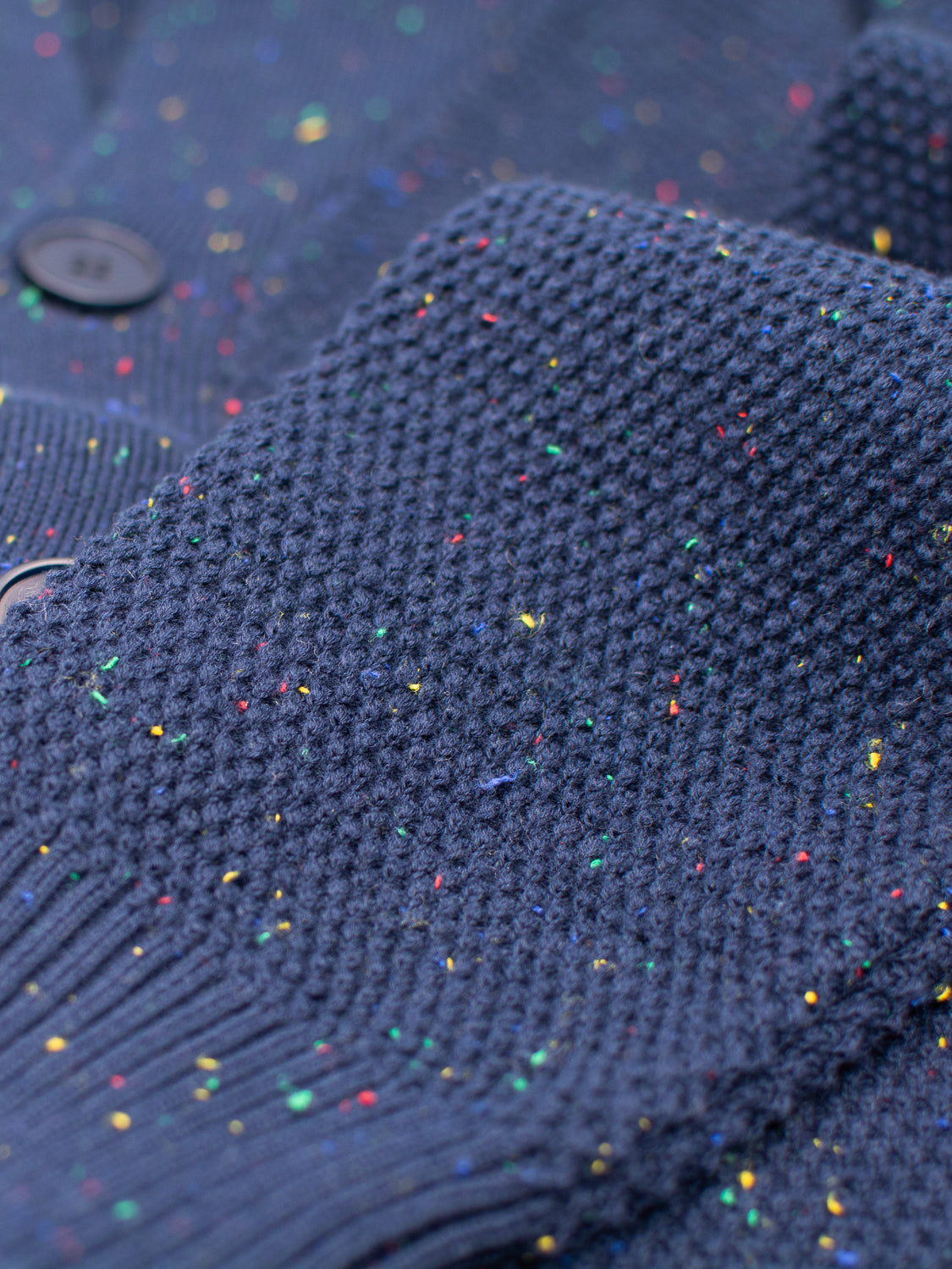 Close-up of a blue, textured Vermont Shawl Collar Cardigan by ID Menswear, crafted from speckled yarn with multicolored specks and a waffle pattern. Visible buttons on the left edge enhance its cozy sweater design.