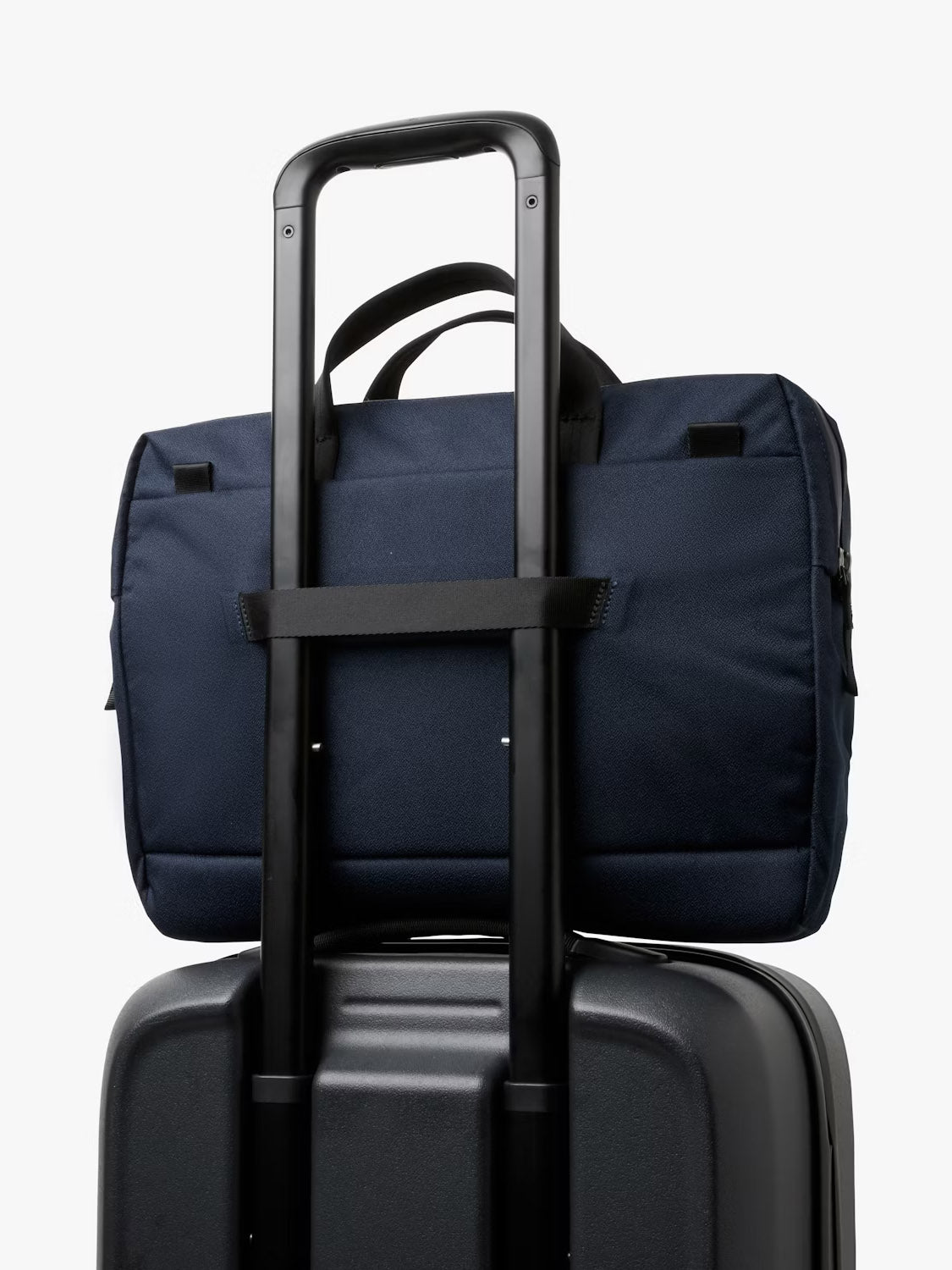 The Bellroy Via Work Bag - 14L in navy blue, featuring a padded laptop sleeve, is strapped to the handle of a black rolling suitcase against a plain white background.