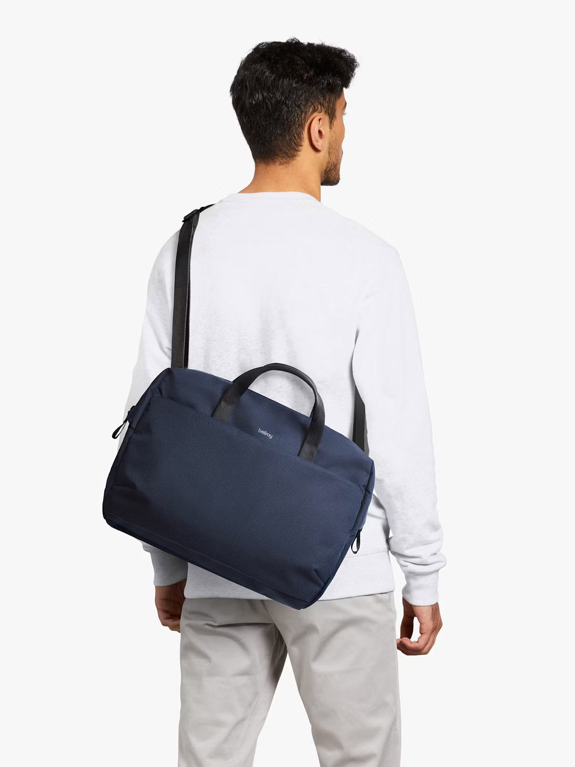 A man in a white long-sleeve shirt and light gray pants stands facing away, carrying the Bellroy Via Work Bag - 14L in navy blue, featuring black handles, a black shoulder strap, and a padded laptop sleeve.