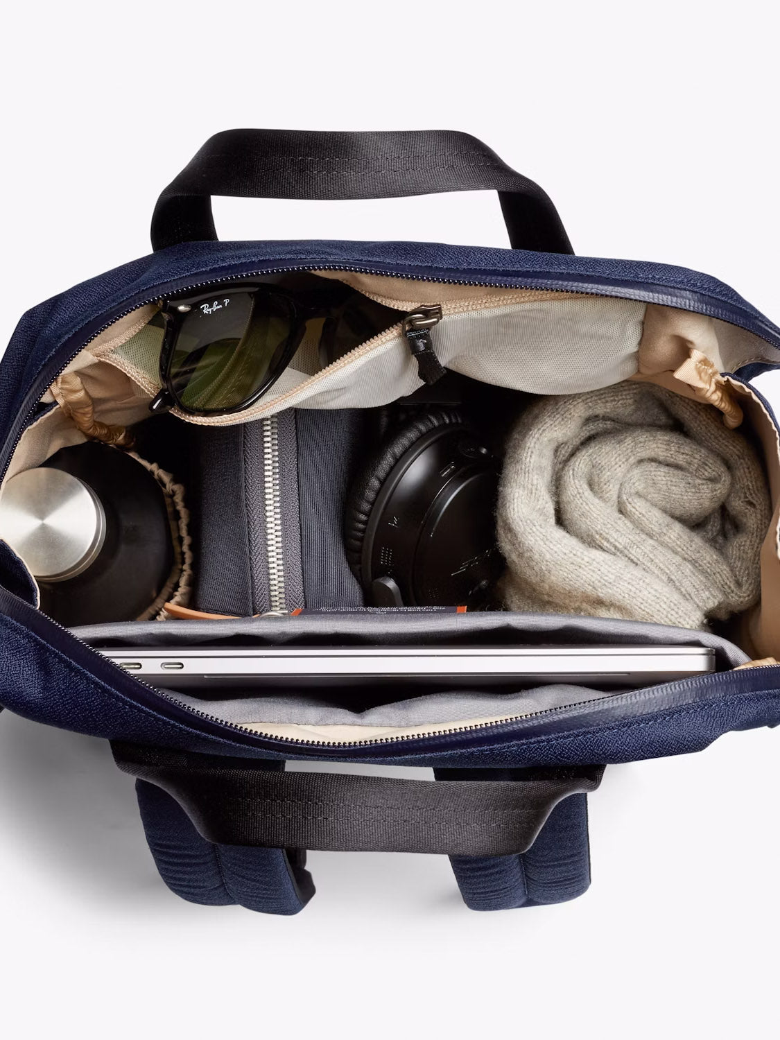 A top view of the open Bellroy Via Workpack - 18L in navy reveals neatly arranged items: sunglasses, water bottle, zip pouch, headphones, a rolled-up grey sweater, and a laptop in its padded sleeve—ideal for commuters.