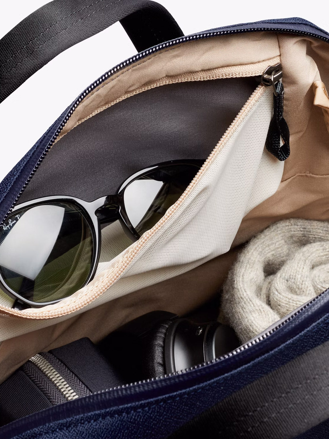 The Bellroy Via Workpack - 18L is shown open, displaying its water-resistant zippered design with organized contents—black sunglasses in a mesh pocket, rolled gray knit, camera lens, and zippered pouch—ideal for commutes or use as a laptop backpack.