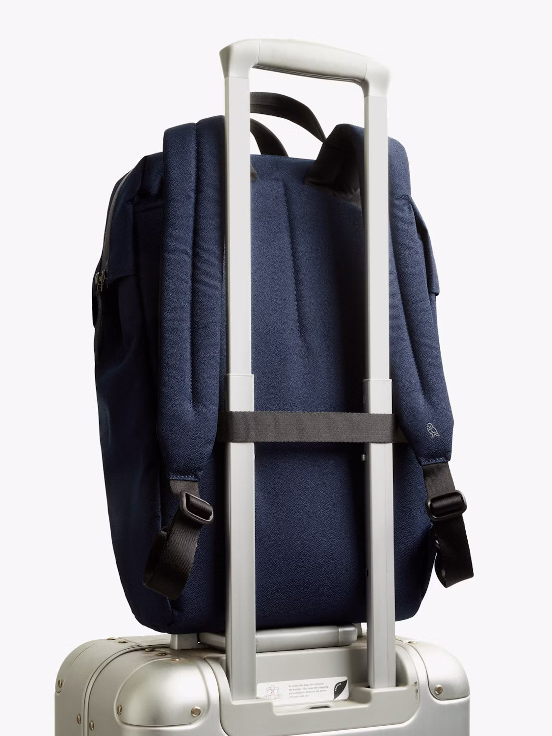 The Bellroy Via Workpack - 18L, a navy blue water-resistant laptop backpack, is attached to the handle of a silver rolling suitcase with its back strap, set against a plain white background.