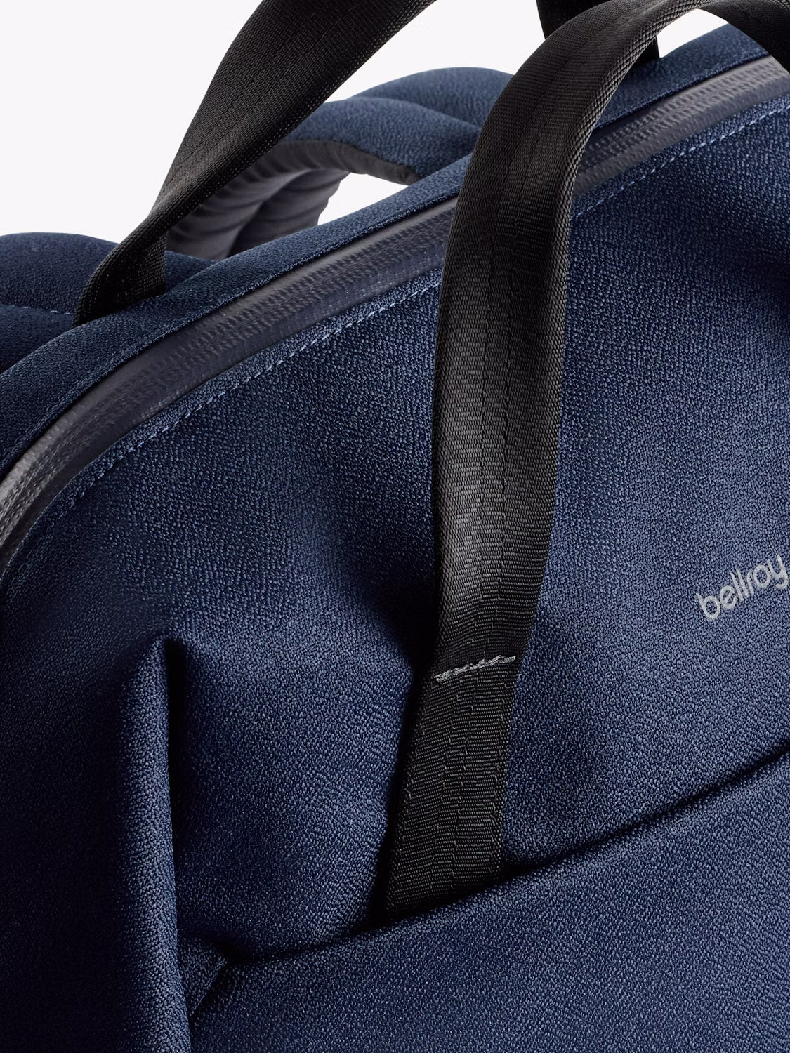 Close-up of the top section of the Bellroy Via Workpack - 18L in navy blue, highlighting its textured water-resistant fabric, black handles, partial zipper, and the visible Bellroy logo on the surface.