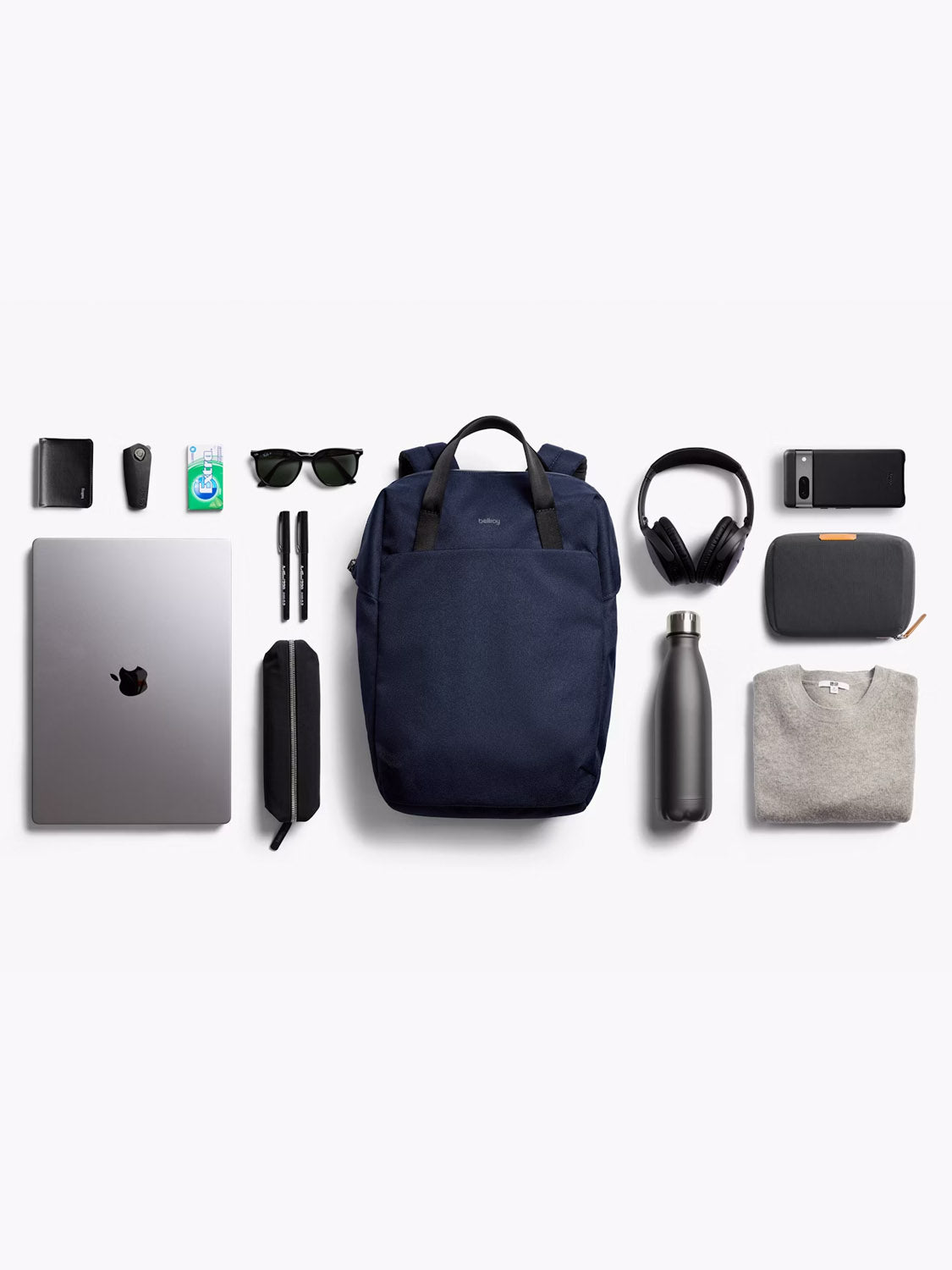 A flat lay of everyday items, including the Bellroy Via Workpack - 18L in navy, displayed with a laptop, sweater, water bottle, sunglasses, pens, headphones, phone, charger, wallet, gum, and a small pouch on a white background.