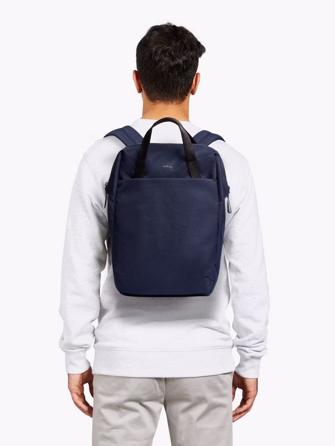 A person in a light gray sweatshirt and beige pants is shown from the back, carrying the Bellroy Via Workpack - 18L, a navy blue water-resistant laptop backpack with black straps, against a plain white background.