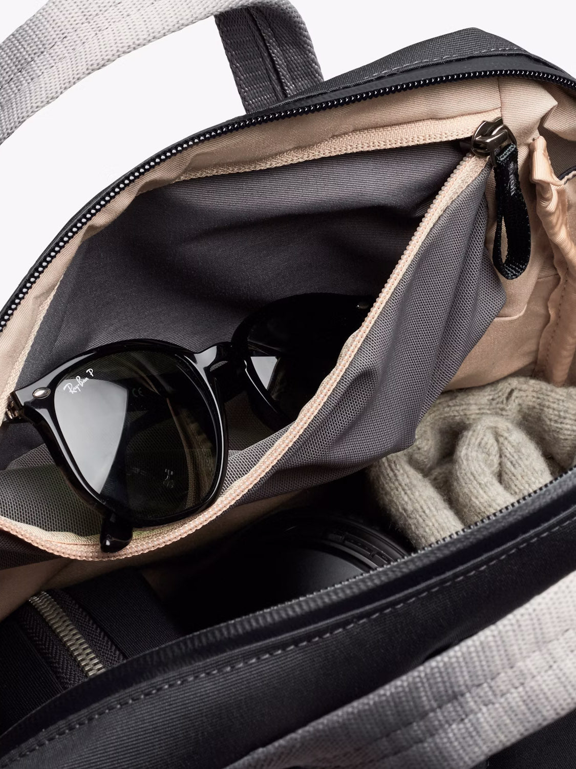 A close-up of an open Bellroy Via Workpack - 18L shows black Ray-Ban sunglasses in the inner pocket, with a knit sweater and other items partially visible inside this water-resistant commuter bag.