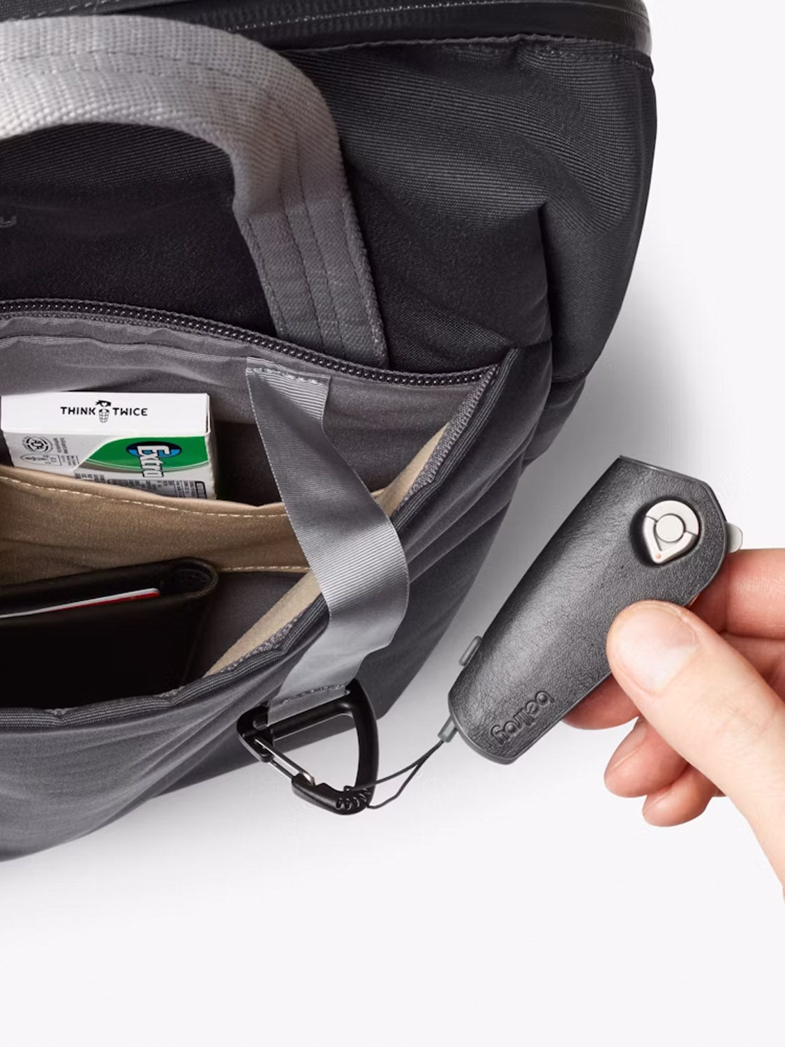 A hand holds a small black gadget connected by a cable to the Bellroy Via Workpack - 18L commuter bag, which has an open pocket containing cards and a wallet.