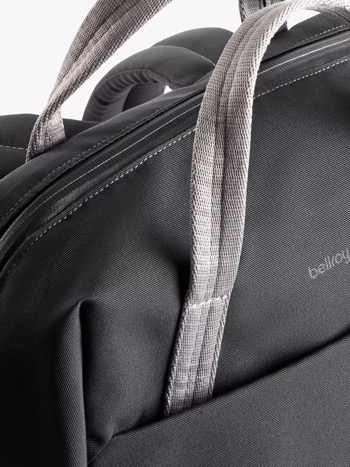 Close-up of the Bellroy Via Workpack - 18L, a black, water-resistant laptop backpack featuring light gray textured handles, visible stitching, and a subtly embossed Bellroy logo near the top right corner.