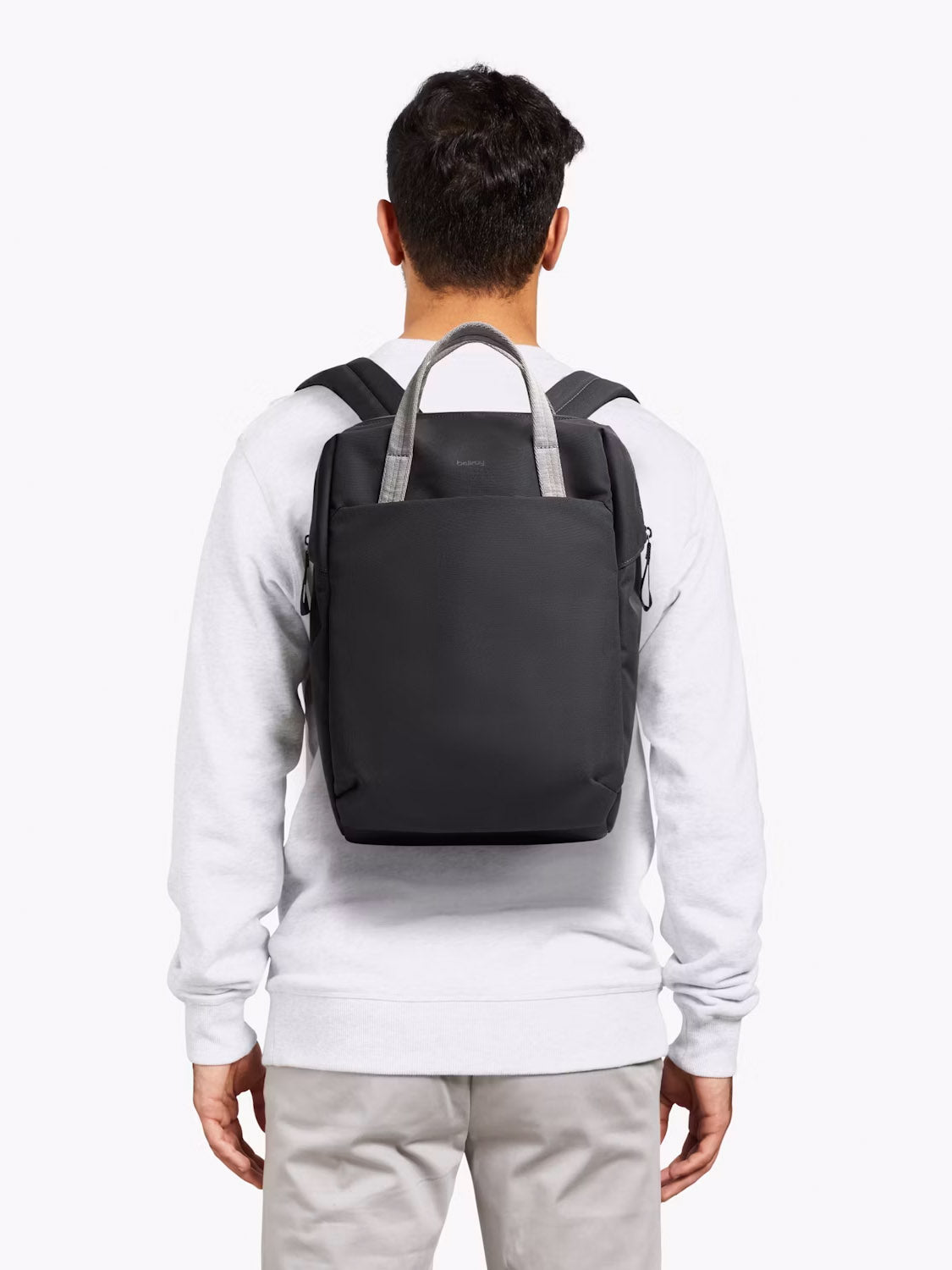 A person is seen from behind in a light gray sweatshirt and beige pants, carrying the Bellroy Via Workpack - 18L, a water-resistant laptop backpack with gray handles. The background is plain white.