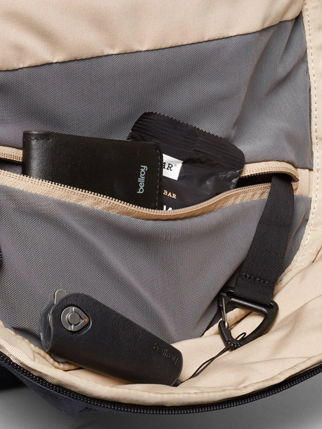 A close-up of the Via Backpack - 20L by Bellroy shows its inside pocket neatly organizing a black wallet, snack bar, and black electronic car key—each arranged neatly beside the laptop sleeve.