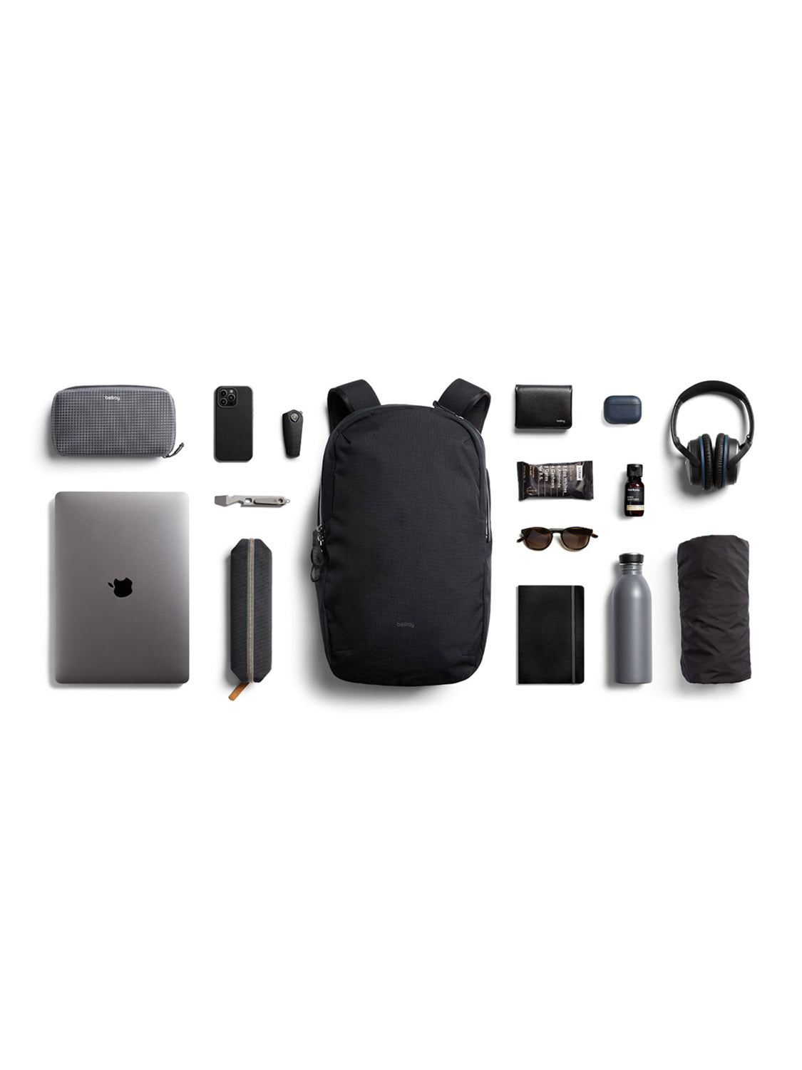 The Bellroy Via Backpack - 20L, a black minimalist bag made from recycled fabric, is displayed with neatly arranged everyday essentials: laptop (in sleeve), phone, wallet, headphones, glasses, bottle, umbrella, notebook, pen, pouch, speaker, cords & sanitizer.