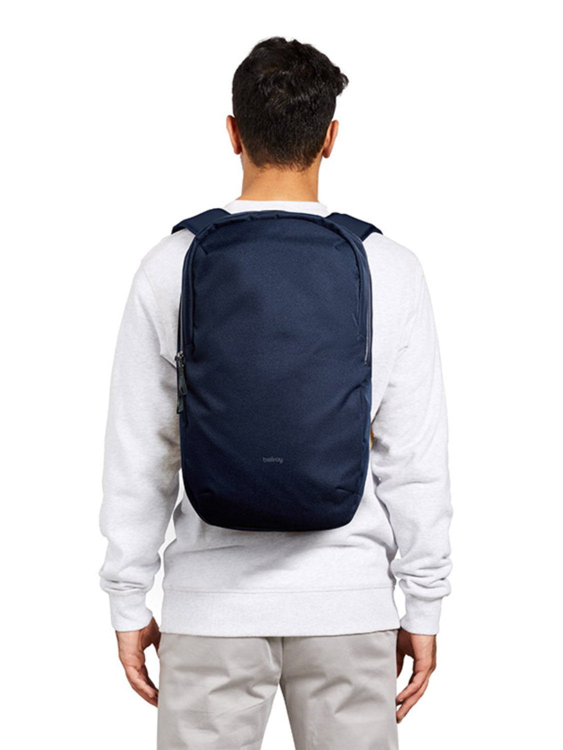 A person in a white sweatshirt and light pants is seen from behind carrying the Bellroy Via Backpack - 20L, which features padded straps and a laptop sleeve. The background is plain white.