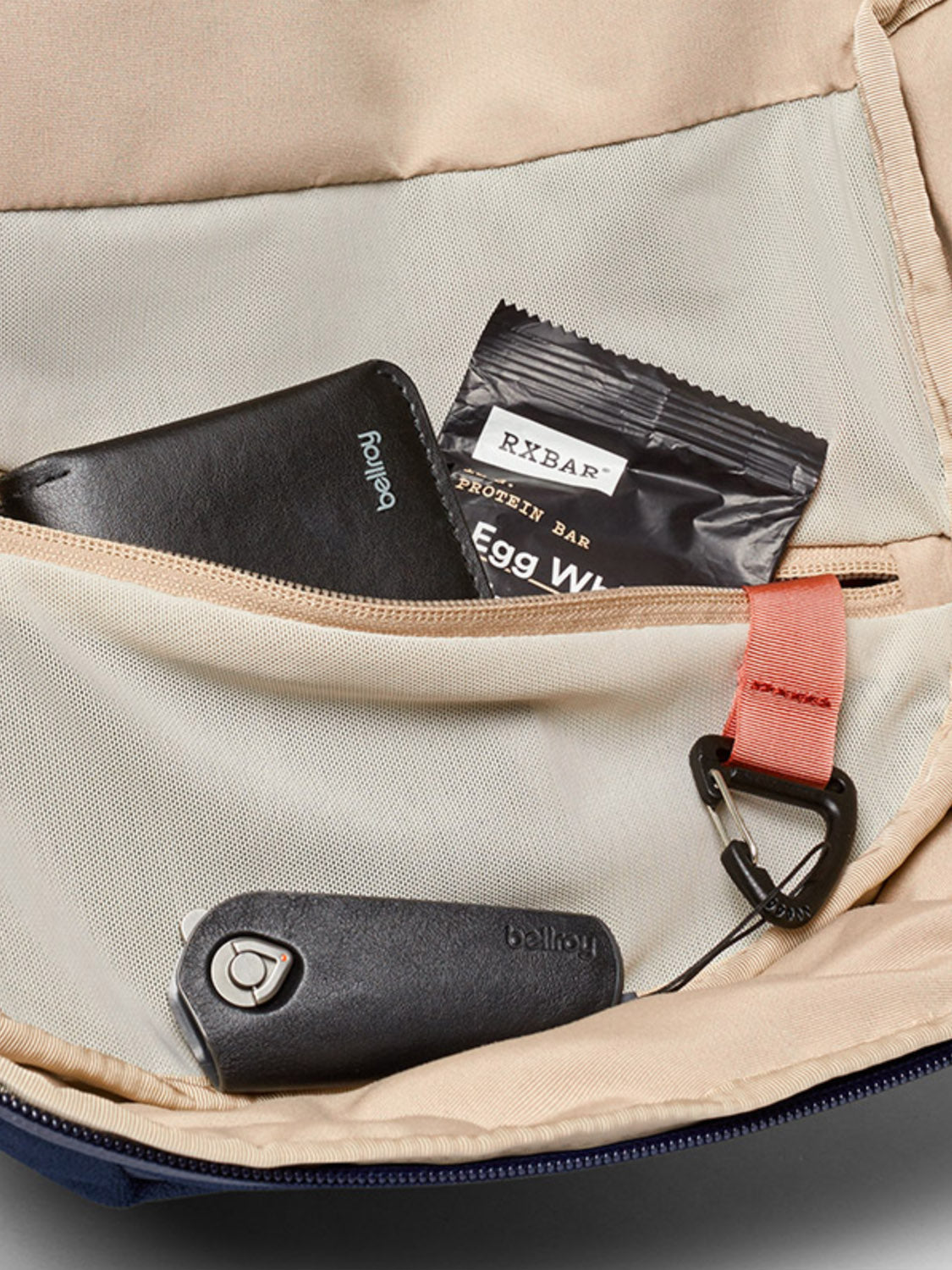A beige Bellroy Via Backpack - 20L with an open front pocket holds a black wallet, a black RXBAR protein bar, a key fob, and a black carabiner clipped to a pink fabric loop.