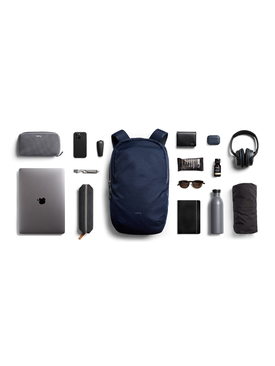 The Bellroy Via Backpack - 20L, shown in navy and crafted from recycled fabric, sits among travel essentials—laptop, water bottle, headphones, sunglasses, phone, and charger. Features include a secure laptop sleeve and organized compartments on white backdrop.