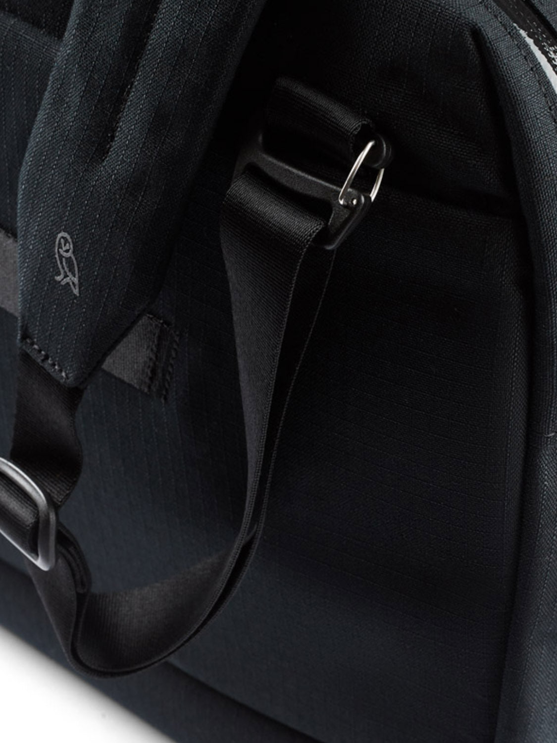 Close-up of the Bellroy Via Work Bag - 14L in black, showing a shoulder strap with metal hardware and a small embroidered owl logo. Made from recycled fabric with visible stitching and includes a padded laptop sleeve inside.