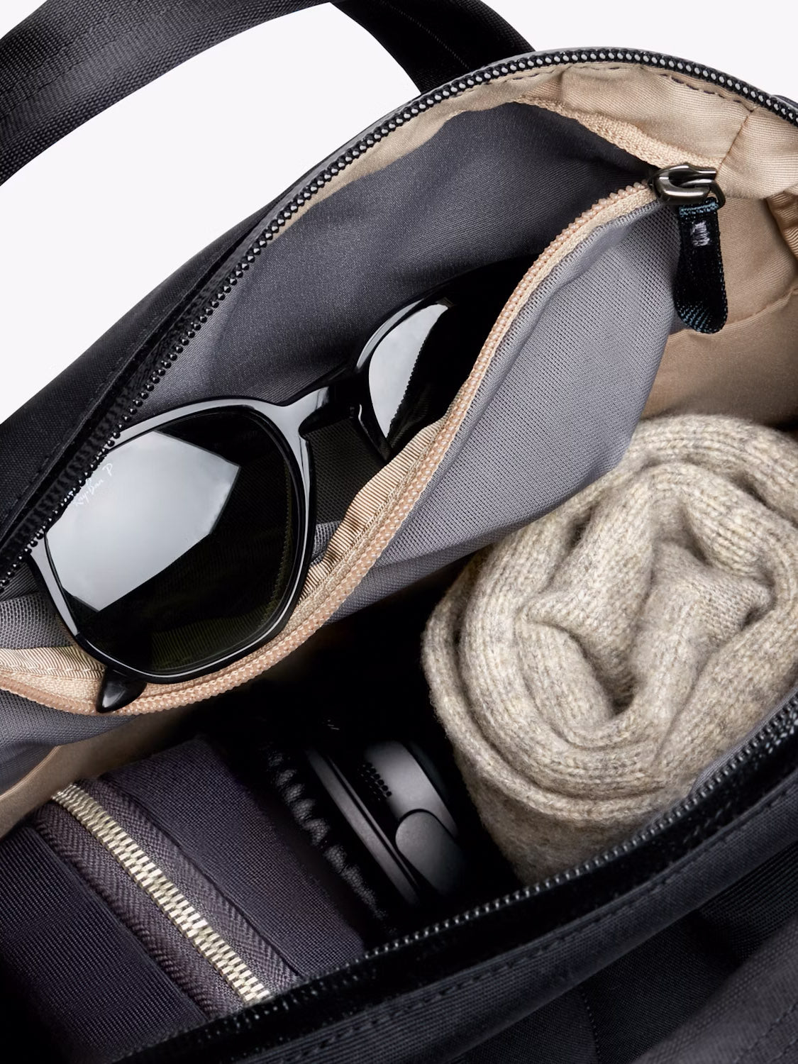 A close-up of the Bellroy Via Workpack - 18L, water-resistant and open, showing black sunglasses in an inner pocket, a rolled beige sweater, a striped fabric item, and a black cylindrical object inside.