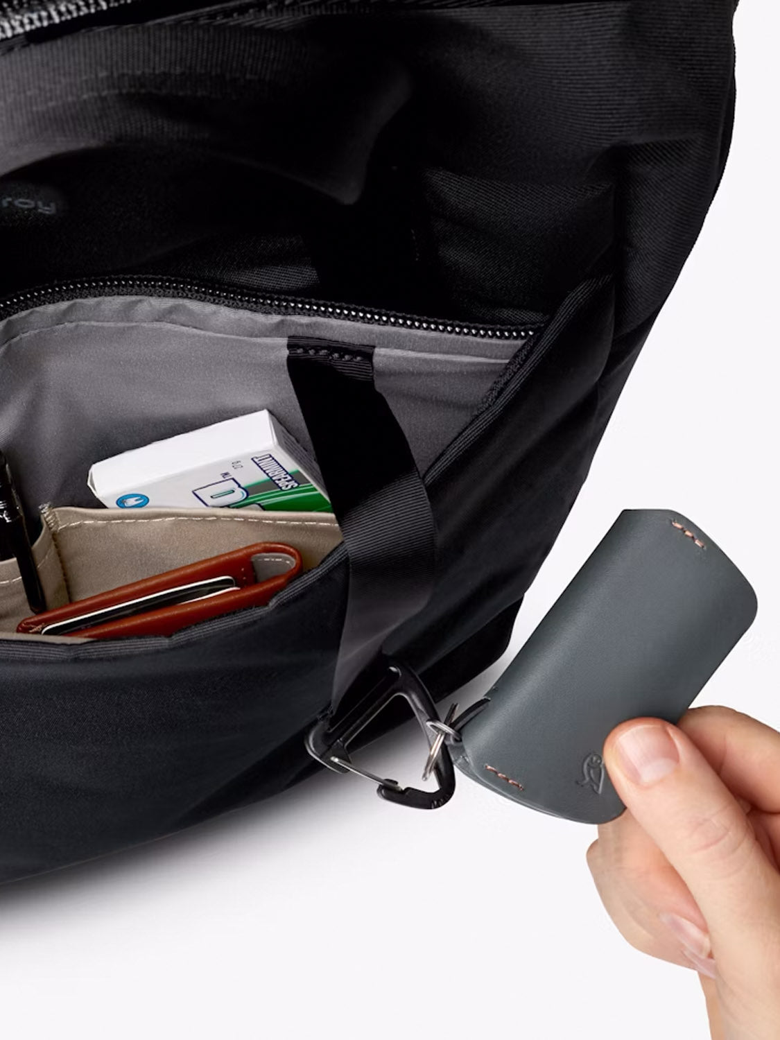 A hand clips a small, grey device to the key ring of a Bellroy Via Workpack - 18L. The open black bag displays a pen, phone, and tissues inside its organized compartment, highlighting its design for everyday commuting.