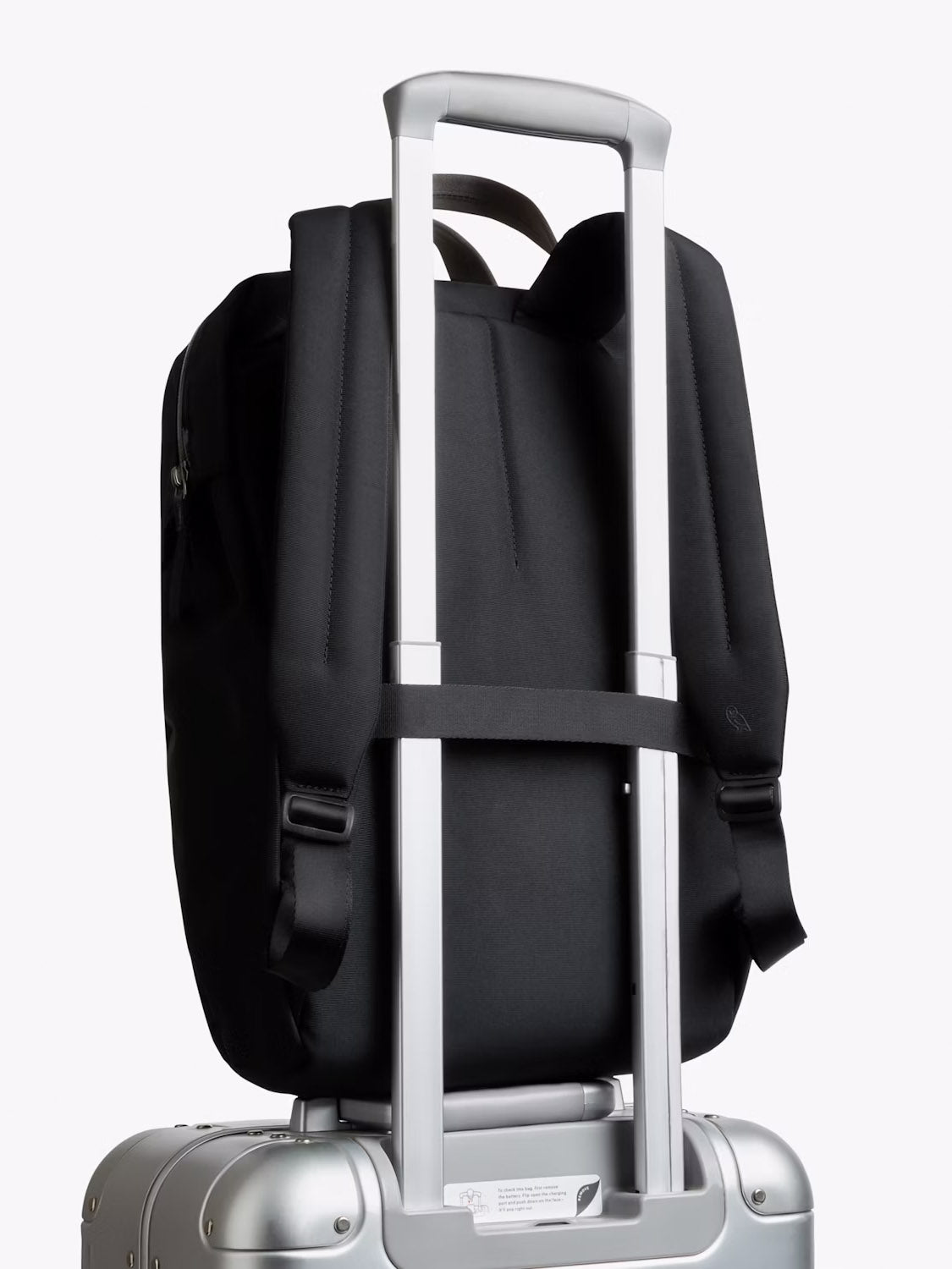 The Bellroy Via Workpack - 18L is securely attached to the extended handle of a silver rolling suitcase, with its straps facing outward against a plain white background.