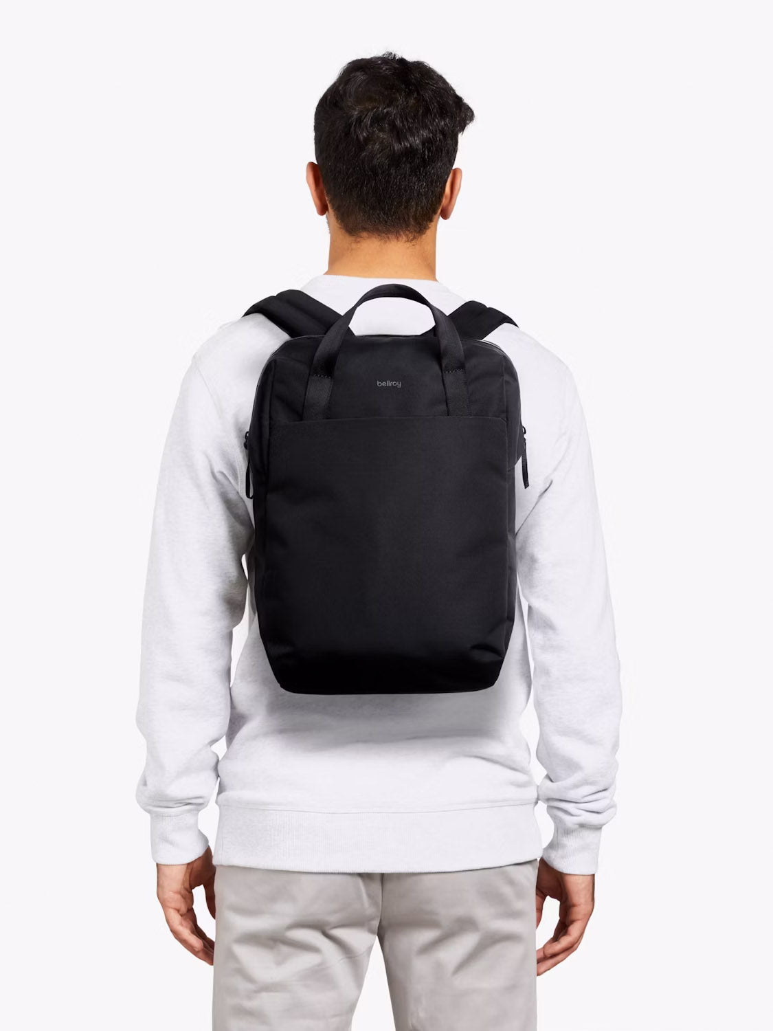 A person in a white sweatshirt and light beige pants, seen from behind, carries the Bellroy Via Workpack - 18L, a black water-resistant laptop backpack with short handles and shoulder straps, against a plain white background.
