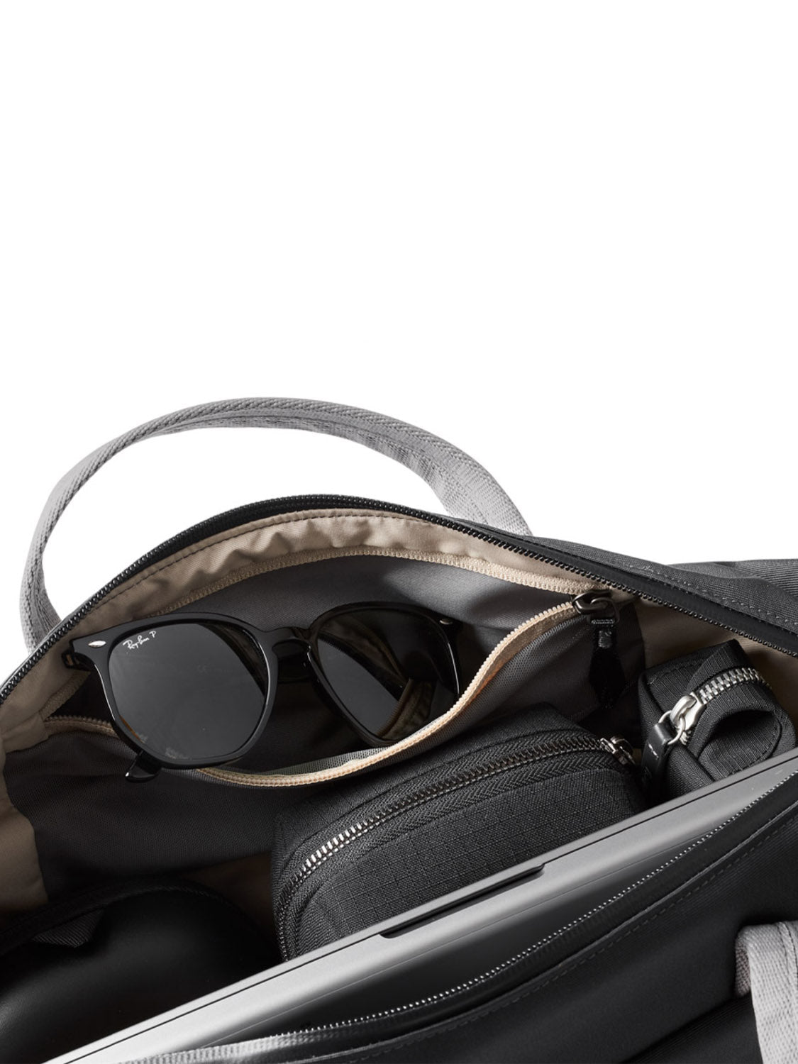 A close-up of the Bellroy Via Work Bag - 14L in black with gray handles, open to show its main compartment containing black sunglasses, a zippered pouch, and part of a laptop or tablet.