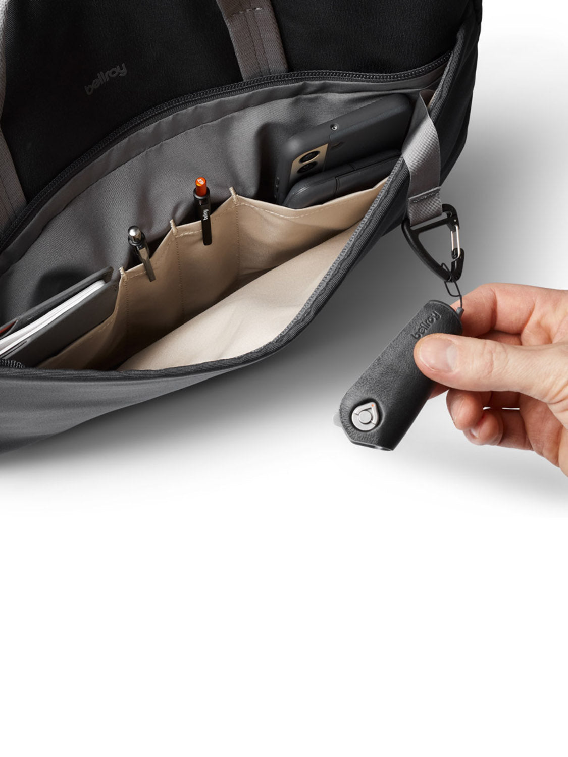 A hand removes a black key holder from a hook inside a Bellroy Via Work Bag - 14L, with organized pockets holding pens, a phone, and other essentials.