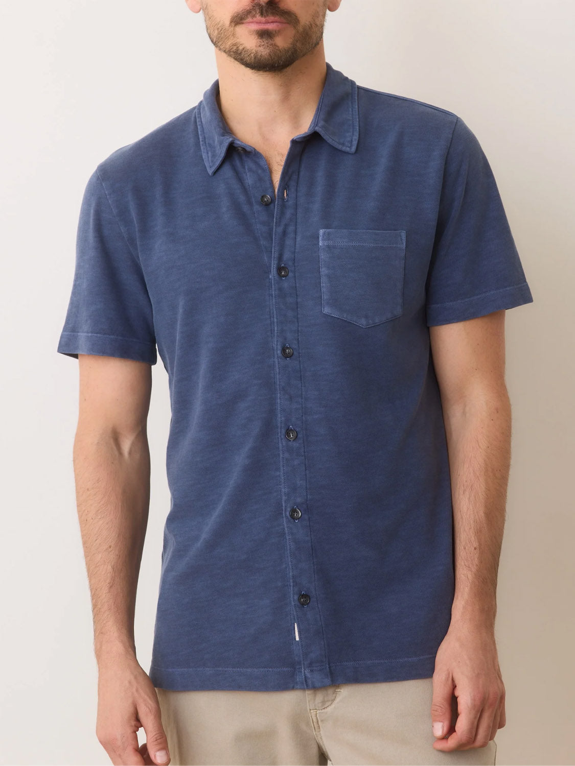 A man wearing Marine Layers navy Vintage Heavy Slub Button Down and beige pants stands against an off-white background, showcasing the shirts casual garment-dyed, slightly faded vibe.