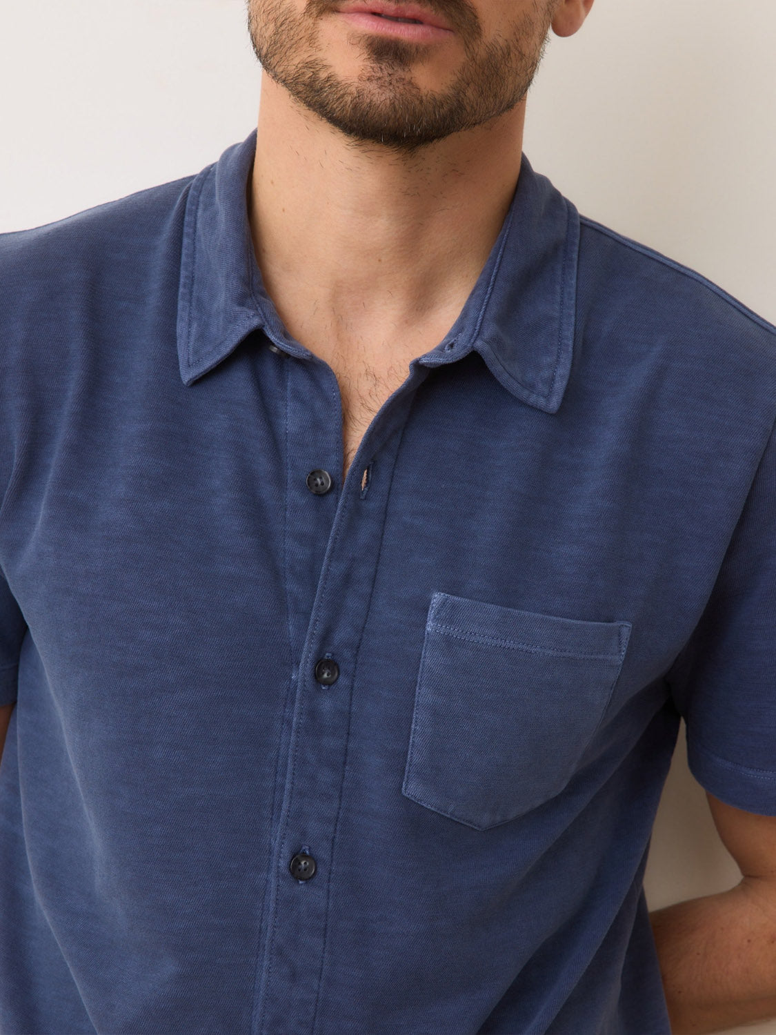 A man wearing the Marine Layer Vintage Heavy Slub Button Down in blue, featuring a front pocket, stands against a white wall.