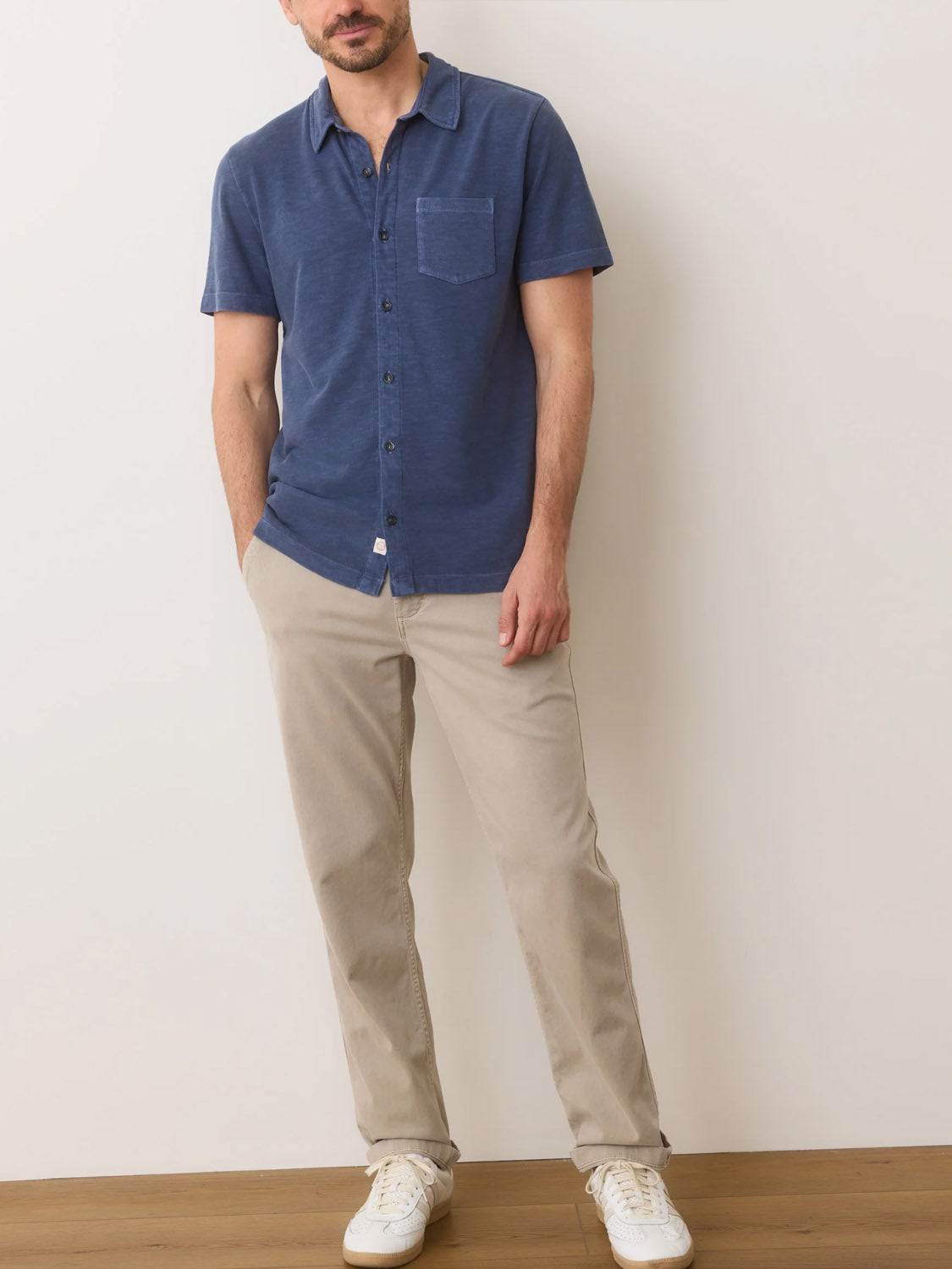 A man stands against a light wall in a Marine Layer Vintage Heavy Slub Button Down, blue with short sleeves and a chest pocket, along with beige pants and white sneakers. His hands are in his pockets as he leans slightly to one side.