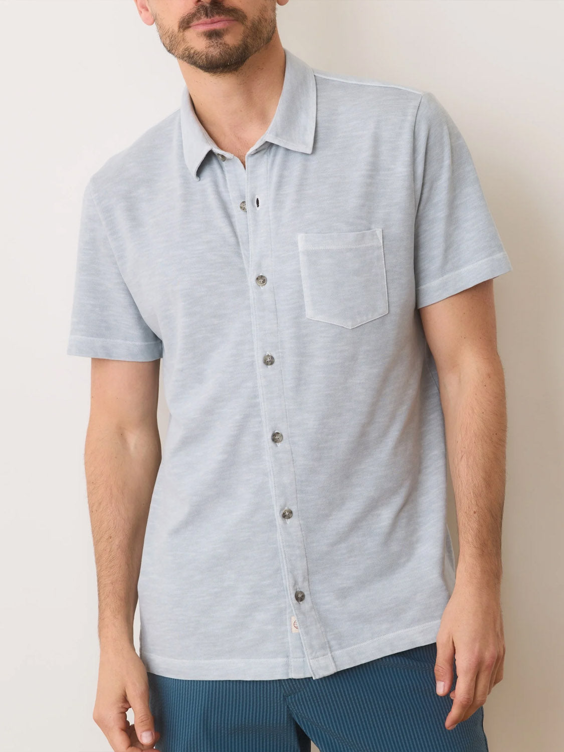 Wearing the Marine Layer Vintage Heavy Slub Button Down, a light blue short-sleeve shirt with a front pocket, they pair it effortlessly with dark blue pants against a plain white background.