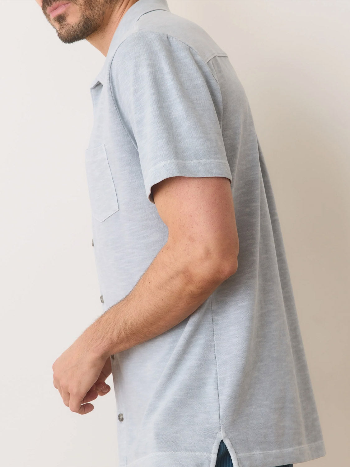 The individual wears a Marine Layer Vintage Heavy Slub Button Down, a light gray, short-sleeve shirt with a chest pocket. The garment-dyed fabric boasts a vintage slub texture. A side view shows part of their face and arm against a plain white background.