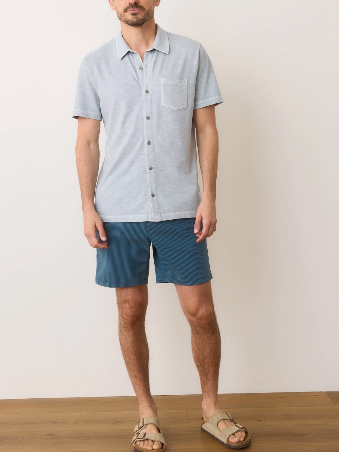 A man stands on a wooden floor against a white wall wearing the Marine Layer Vintage Heavy Slub Button Down in light blue with a chest pocket, dark blue shorts, and light brown sandals.