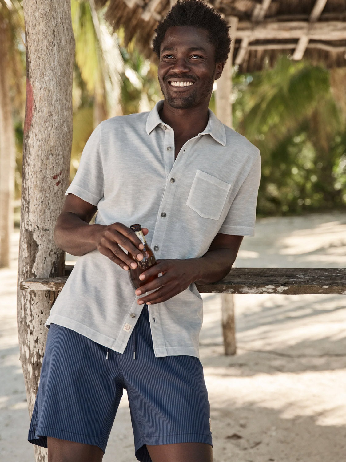 A smiling man in a Marine Layer Vintage Heavy Slub Button Down and blue shorts stands under a thatched structure on the sandy beach, holding a drink. Palm trees and wooden beams are visible in the sunny background.