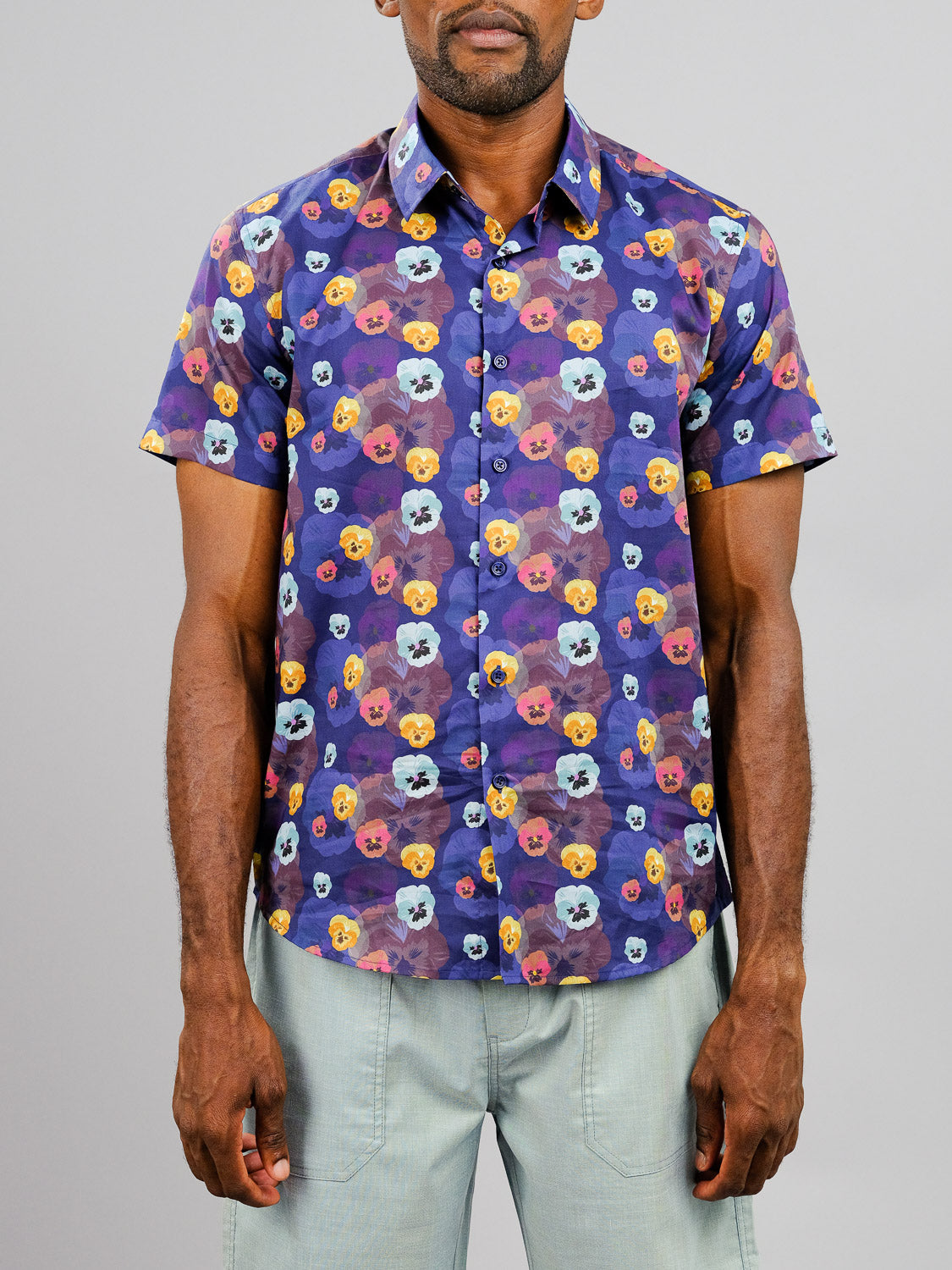 A man wearing the ID Menswear Viola Printed Cotton Shirt, featuring a colorful pansy pattern on dark purple, paired with light pants and standing against a plain gray backdrop.