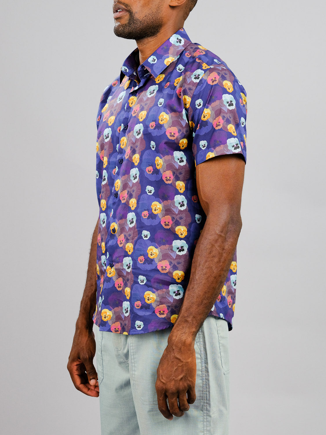 A man wears the Viola Printed Cotton Shirt by ID Menswear, featuring vibrant prints and short sleeves. He stands sideways against a plain gray background, pairing the colorful cotton shirt with light gray pants; his face is partially out of frame.