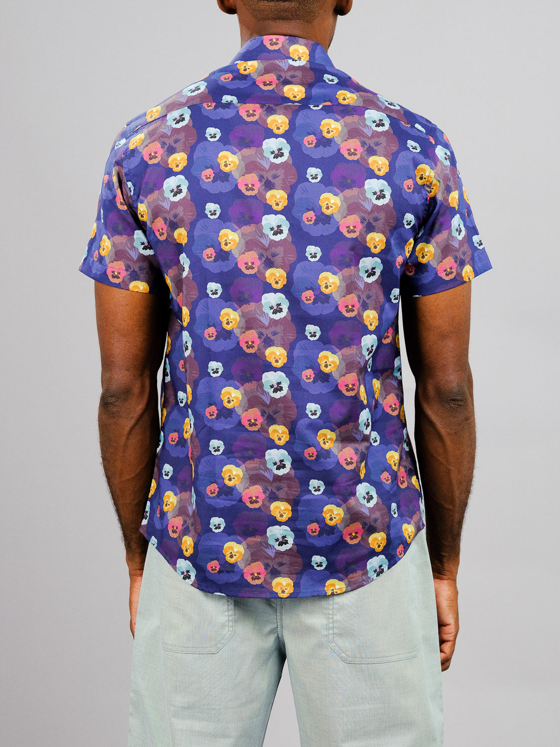 A person faces away wearing the ID Menswear Viola Printed Cotton Shirt, a mainly purple cotton top with vibrant multicolored floral prints, paired with light gray pants.