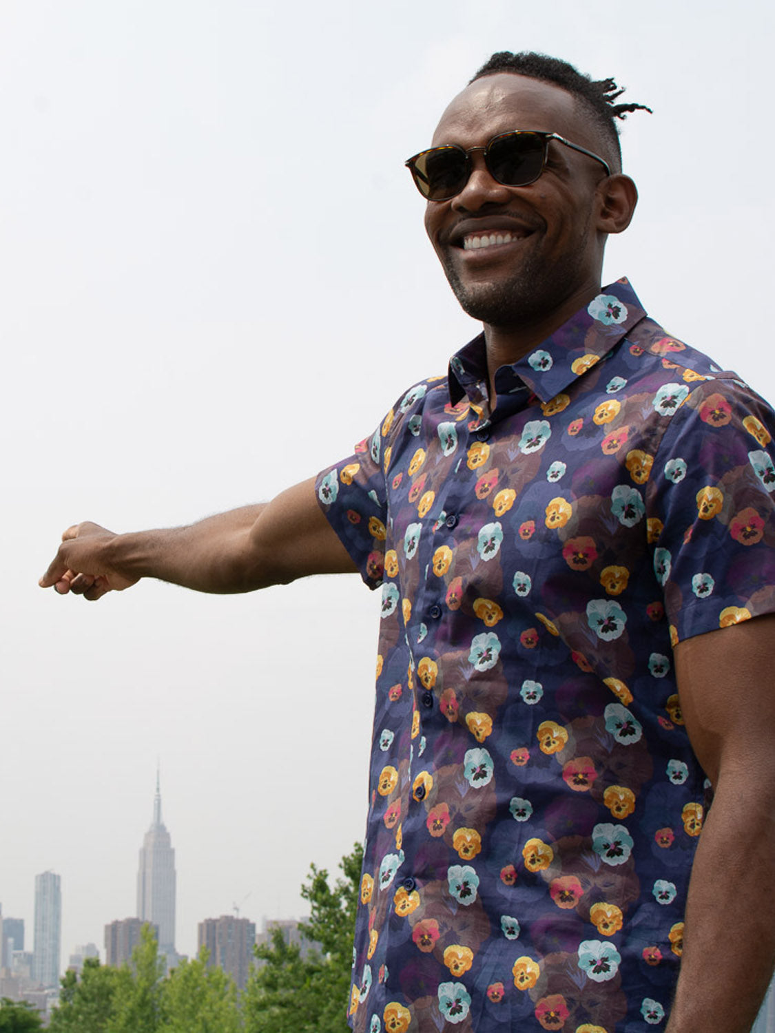 A man in the Viola Printed Cotton Shirt by ID Menswear and sunglasses stands outdoors, smiling as he points toward the Empire State Building on the New York City skyline.