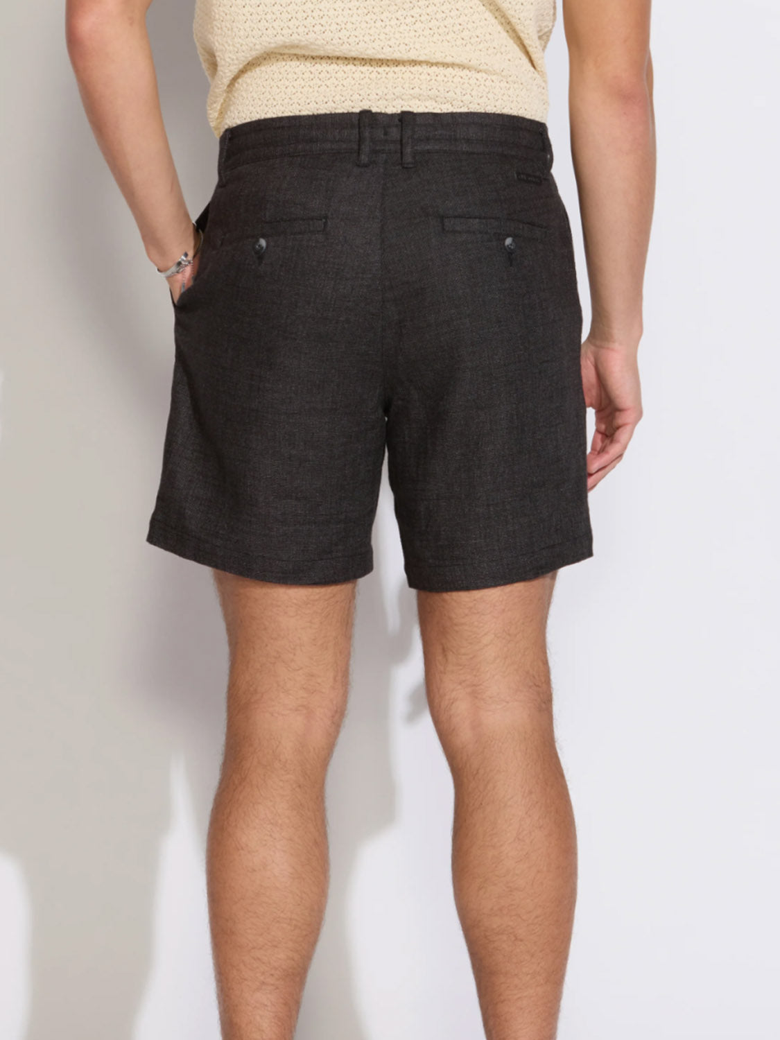 A person is seen from behind wearing Civil Society's Vito Relaxed Fit Short in dark gray linen-blend, featuring belt loops, side and back welt pockets. They pair it with a light beige textured shirt against a plain light background.