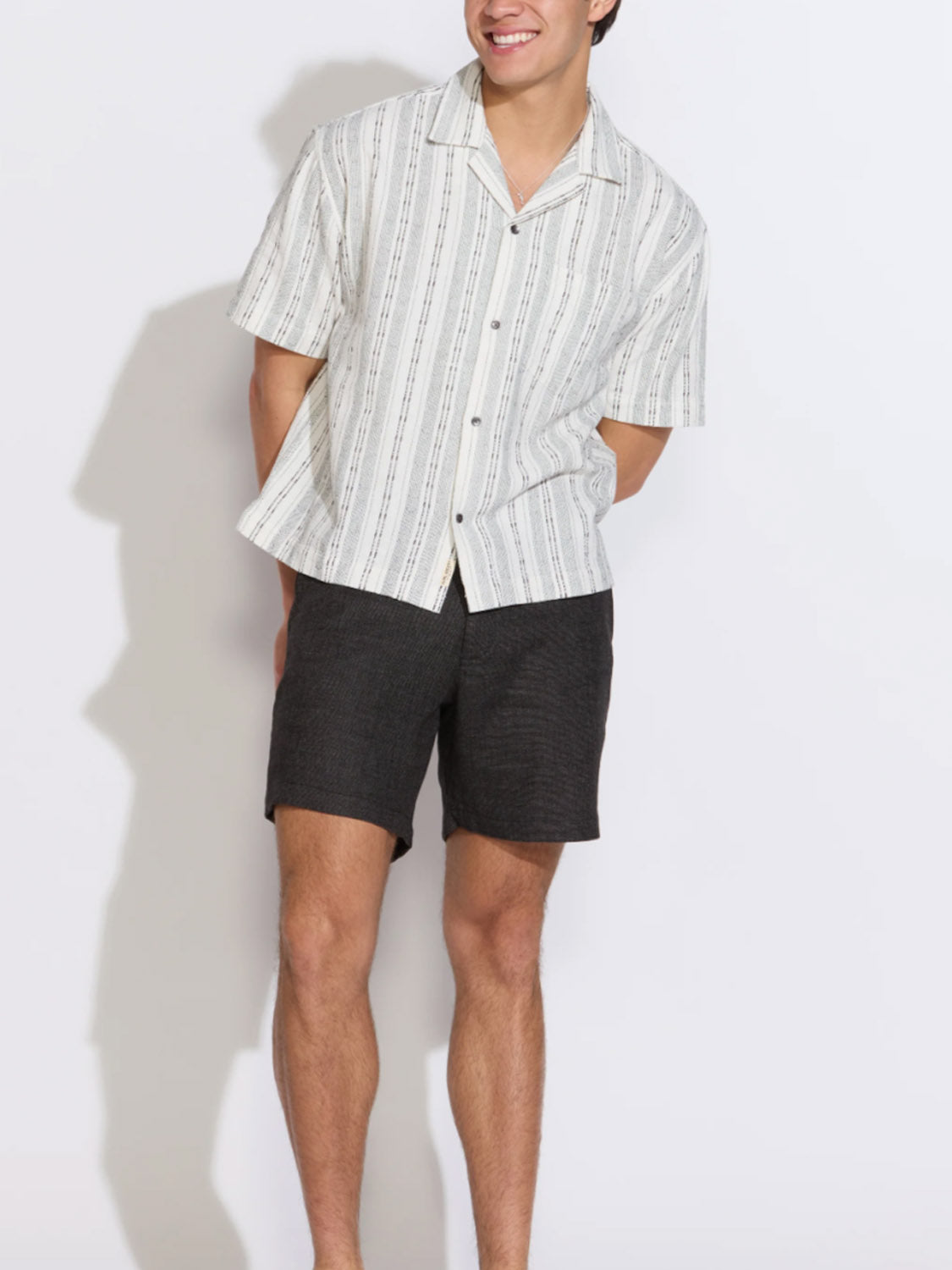 A person smiles with hands in pockets, wearing Civil Society's Vito Relaxed Fit Shorts and a short-sleeve white-and-gray striped button-up shirt, standing against a plain white background.