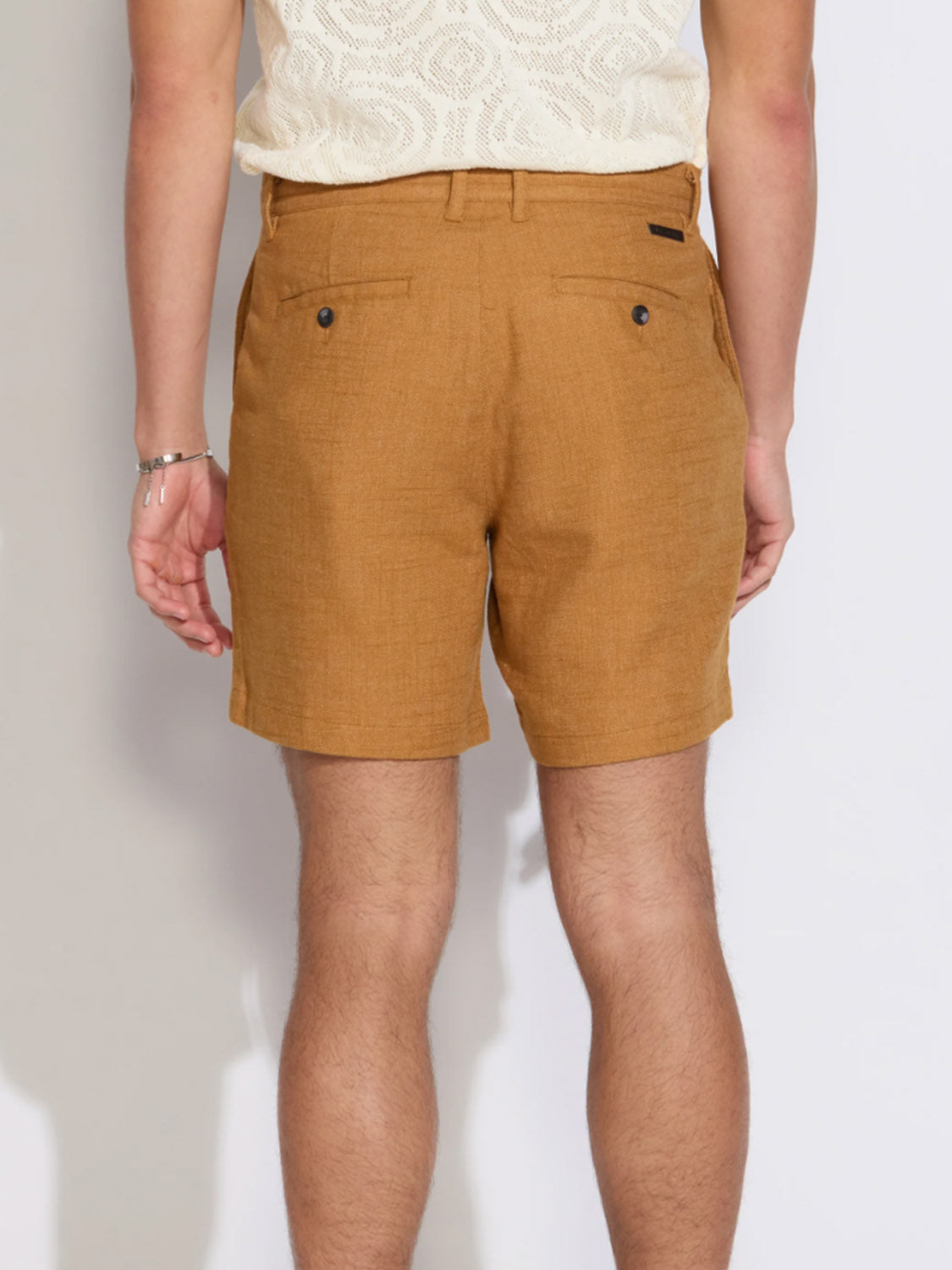 A person stands facing away, showing only their lower back and legs, wearing Civil Society's Vito Relaxed Fit Short in mustard brown linen-blend with belt loops and buttoned back pockets, paired with a white patterned short-sleeve shirt.