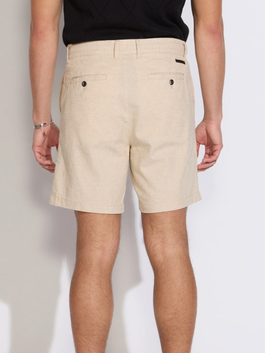 A person is shown from the back, wearing Civil Society’s Vito Relaxed Fit Short in beige—a mid-thigh linen-blend short with belt loops and two buttoned back pockets—paired with a black shirt against a plain white background.